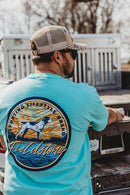 A Lifestyle photo of a man wearing a Fieldstone branded seafoam green short sleeve pocket t-shirt with a custom graphic print of a dog and the sky in the background. The text 'Fieldstone' across the bottom. The front left chest pocket has the fieldstone logo on it.