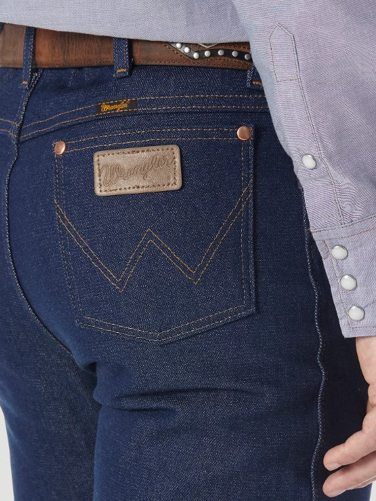 Closeup of back right back pocket of a man wearing Wrangler® Cowboy Cut® slim fit rigid blue jeans with western shirt and belt. Pocket has copper rivets on top corners, "W" stitched on pocket, and neolite brown logo patch.