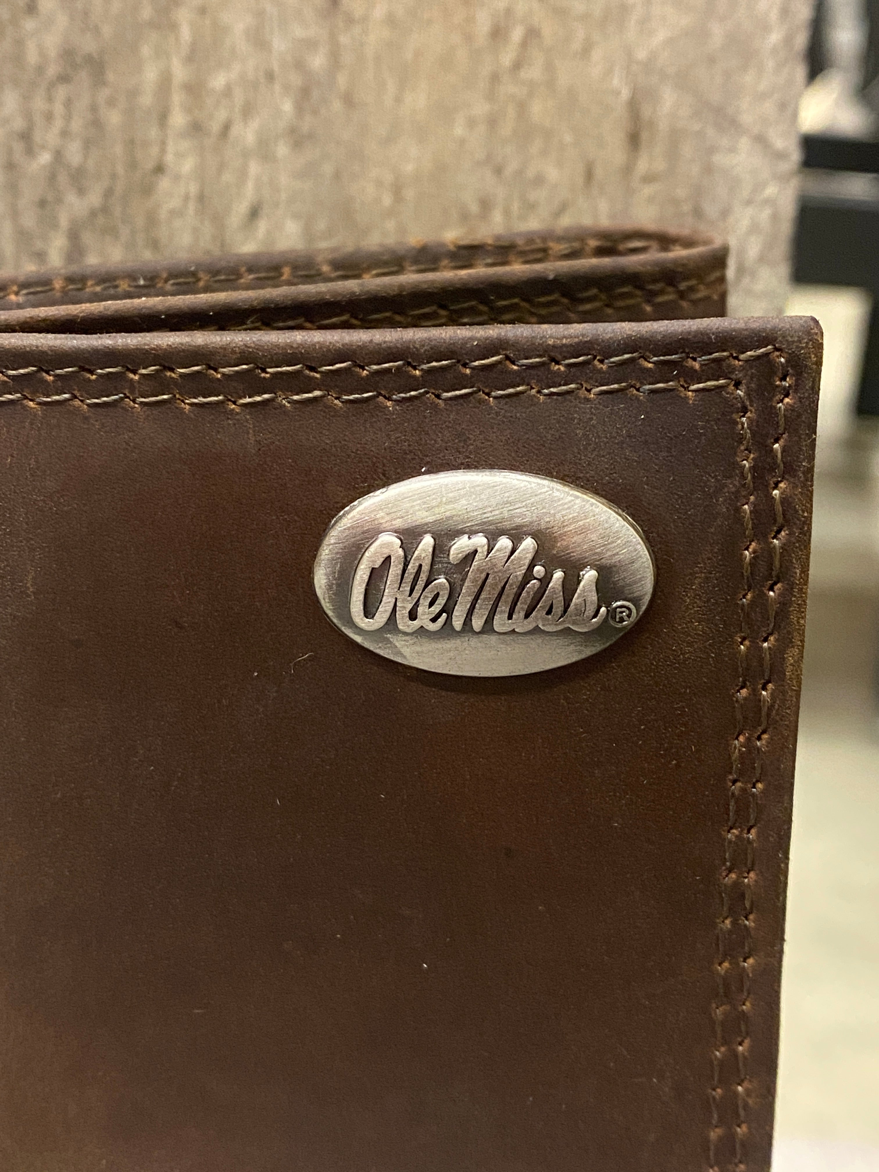 Closeup of the "Ole Miss" logo brass concho on front top right of University of Miississippi "crazy horse" leather tri-fold wallet.